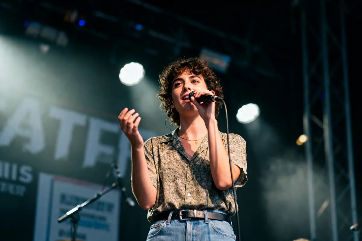 Personne chantant sur scène avec micro, éclairage de concert, chemise à motifs, jean, fond flou de projecteurs.
