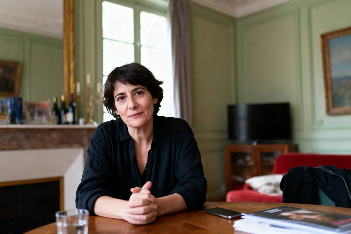 Femme assise à une table, dans un salon élégant avec cheminée et bibliothèque.