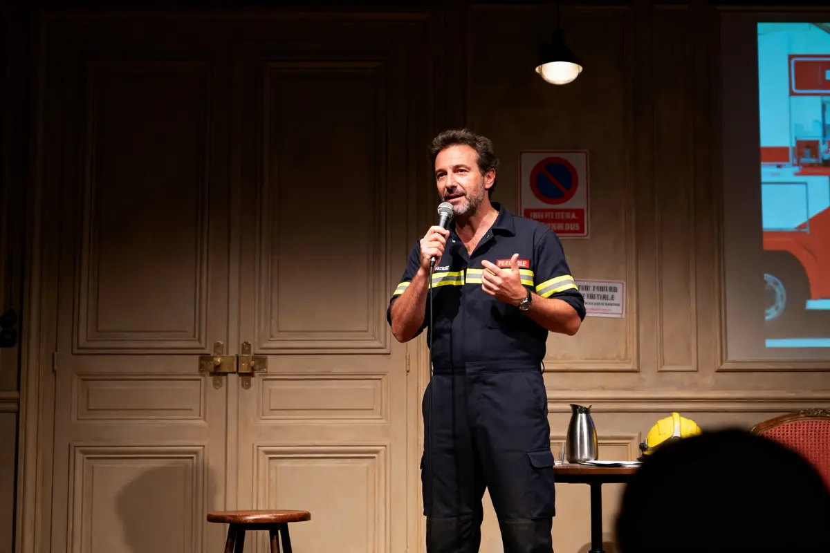 Homme en tenue de pompier sur scène, parlant au micro, fond avec affiches et chaise.