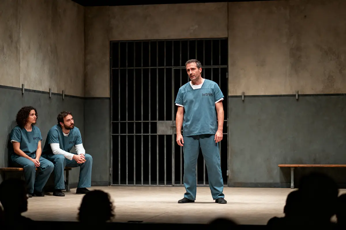 Trois personnages en tenue de prisonniers sur scène, devant une cellule. Salle sombre et austère.