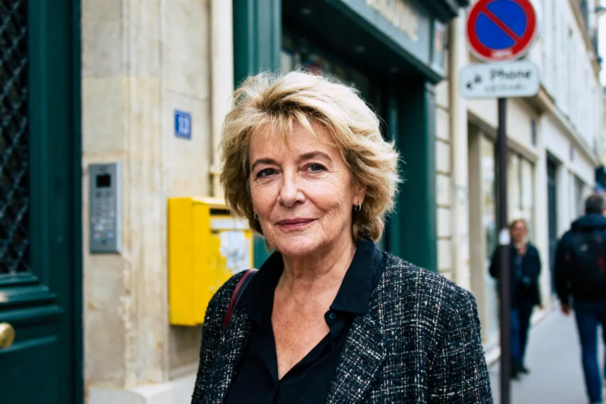 Personne aux cheveux blonds courts, souriante dans une rue devant un mur beige et boîte jaune.