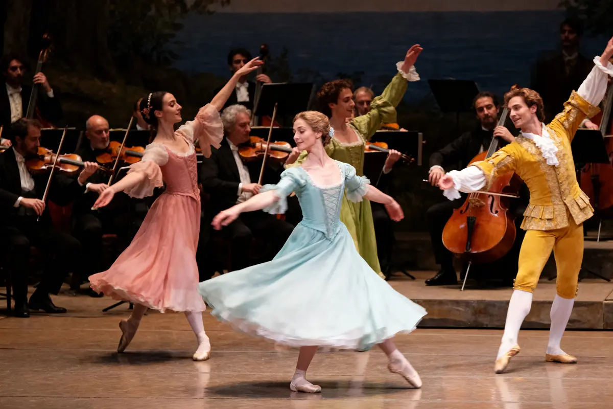 Danseurs en costumes d'époque réalisant un ballet classique, accompagnés par un orchestre en arrière-plan sur scène.