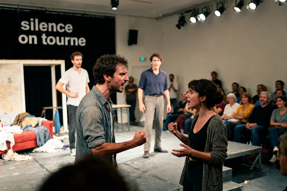 Acteurs sur scène en pleine dispute, public attentif, pancarte "silence on tourne" en arrière-plan.