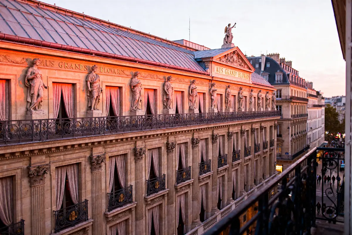 Façade de bâtiment parisien historique au coucher du soleil, statues et balcons. Rue animée en contrebas.